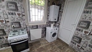 A compact kitchen with a gas stove, a built-in oven, a washing machine, and a radiator. The walls are decorated with heart-shaped cutouts and the word "Home".