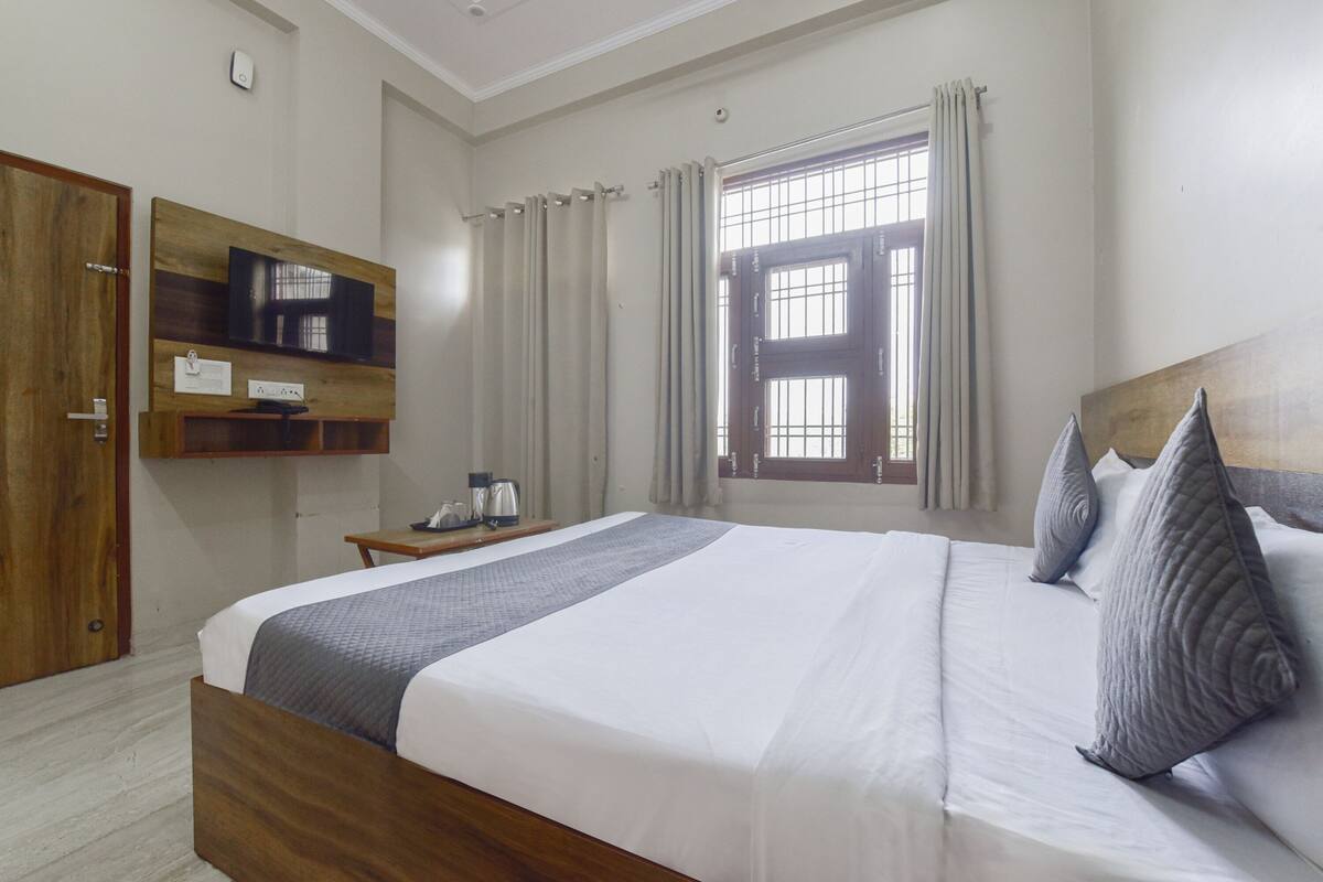 A hotel room with a large bed, a wooden wardrobe, a bedside table with a lamp, a TV mounted on the wall, and a window with curtains.