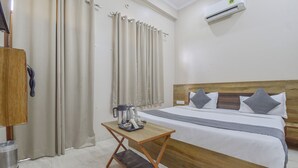 A hotel room with a wooden headboard, a bed with gray and white bedding, a small wooden table with a kettle and cups, beige curtains, and an air conditioning unit on the wall. - Hotel Shivansh (Jaipur)