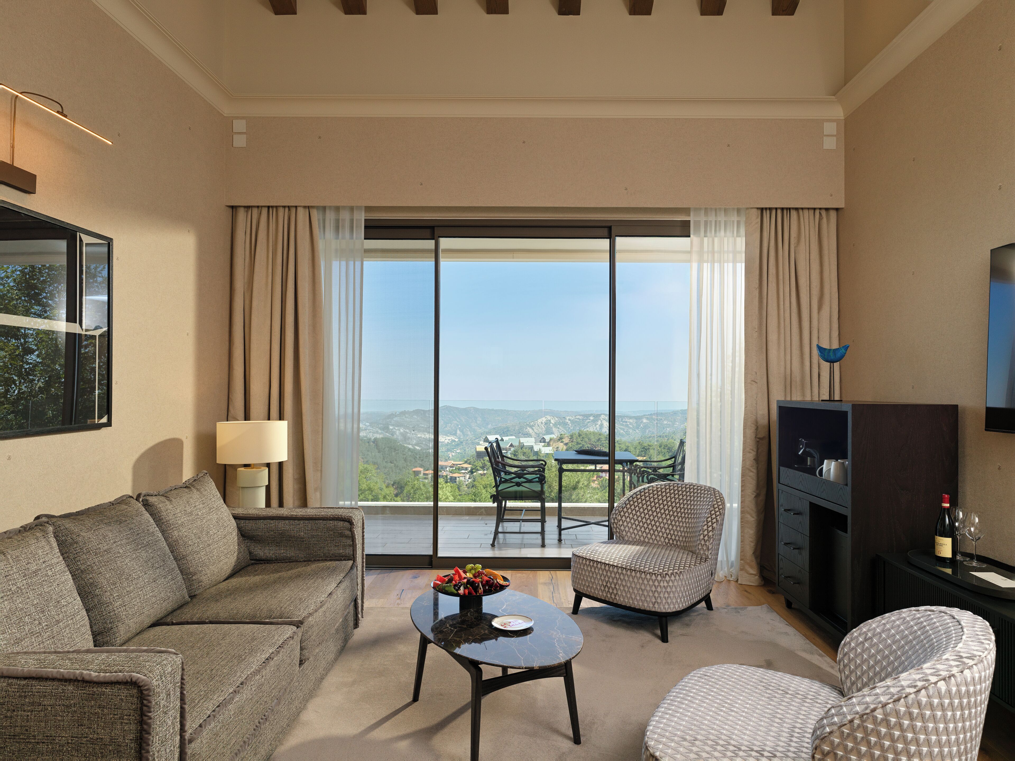 A modern living room with a sofa, a round coffee table, and a TV. There is a balcony with a view of the landscape.