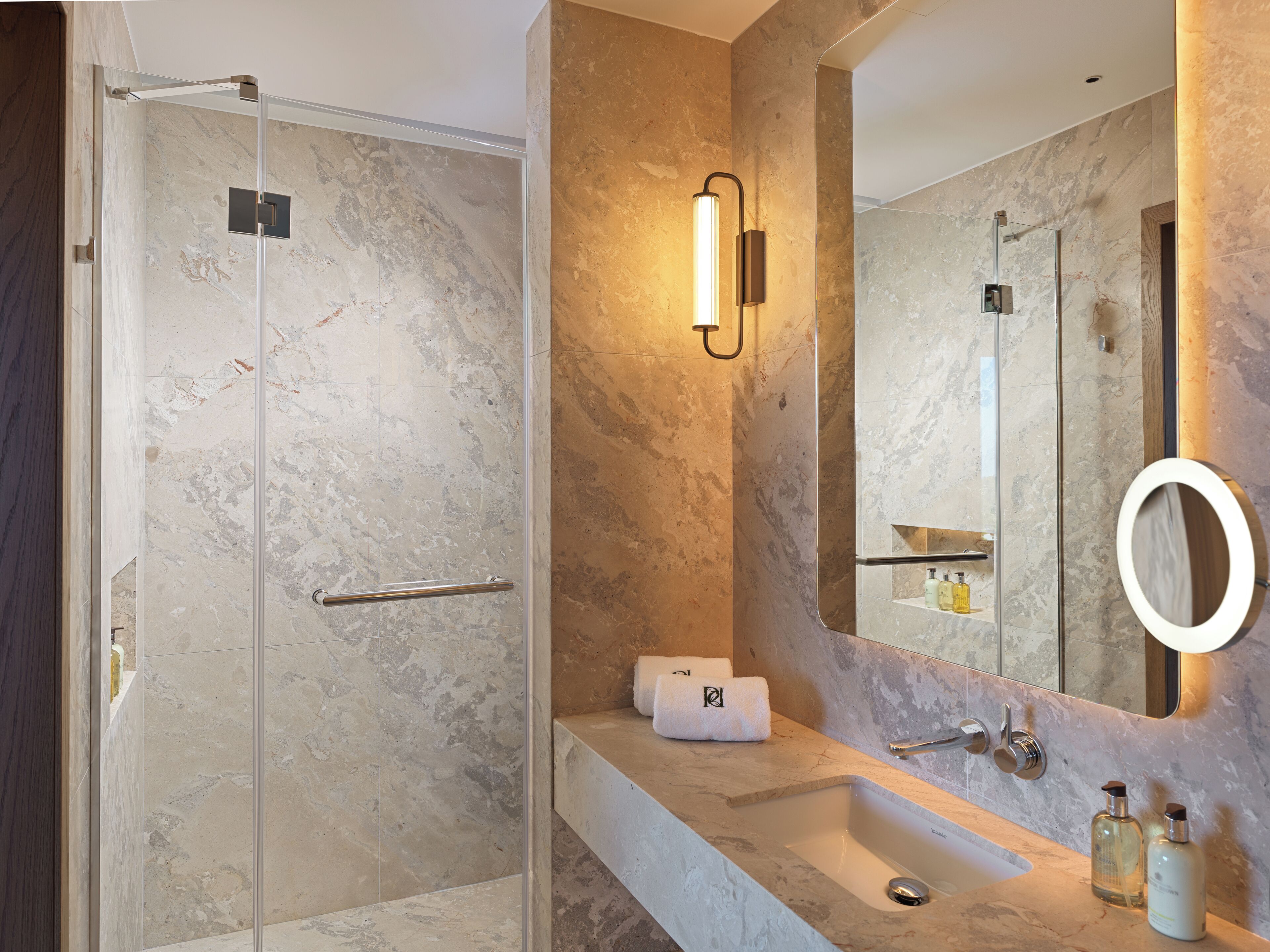 A modern bathroom with a glass-enclosed shower, a marble-tiled wall, a large mirror with a ring light, and a vanity with a sink and toiletries.