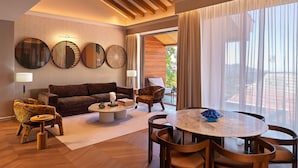 A modern living room with a sofa, armchairs, a coffee table, and a dining table. There are large windows with curtains, a ceiling with wooden beams, and wall-mounted mirrors.