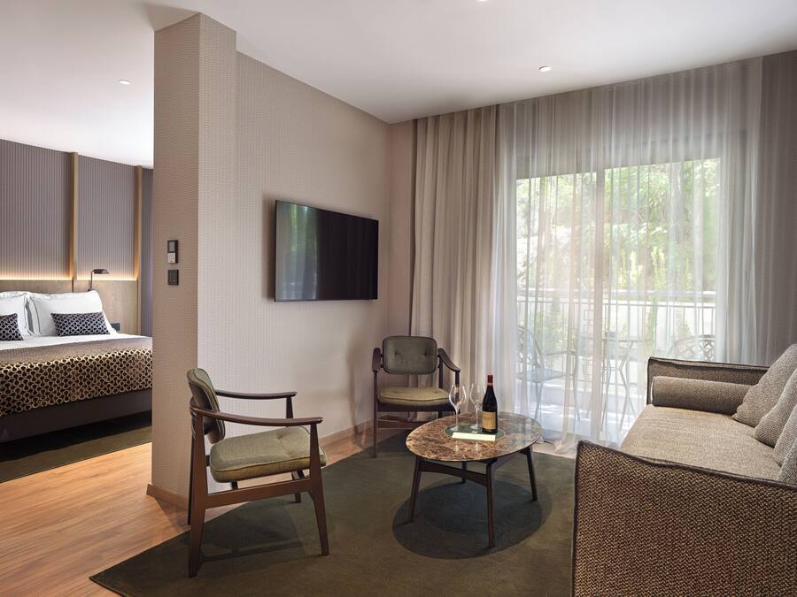Una habitación de hotel moderna con una cama grande, un televisor de pantalla plana, una zona de estar con una mesa y un balcón con cortinas.
