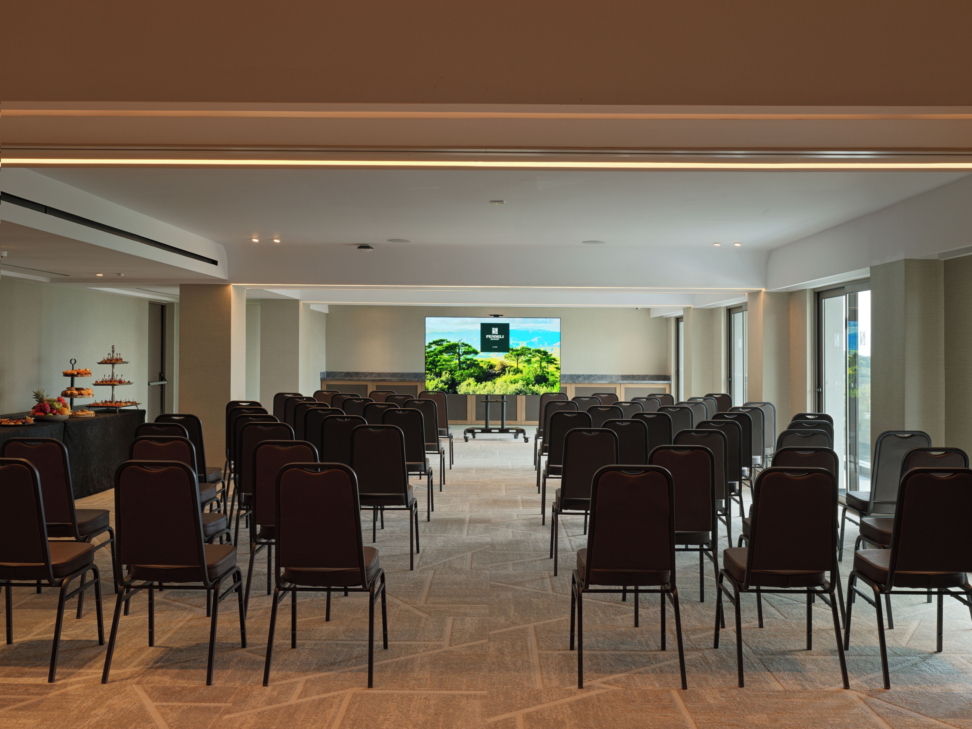 Un salón de conferencias con filas de sillas, una pantalla grande al frente y una mesa de buffet con comida.