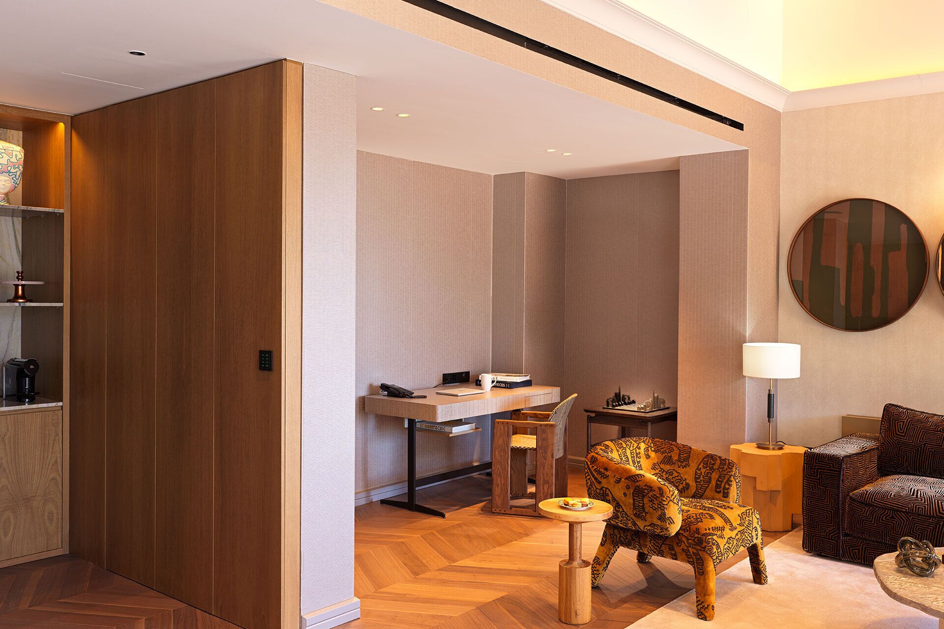 A modern hotel room with a wooden wardrobe, a desk with a chair, a round table, a patterned armchair, and a lamp.