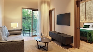A modern hotel room with a sofa, a small table with a bottle and glasses, a TV, and a bed with a green bedspread.