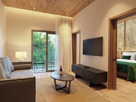 Una habitación de hotel moderna con un sofá, una mesita con una botella y vasos, un televisor y una cama con colcha verde.
