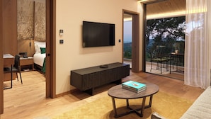 A modern hotel room with a flat-screen TV mounted on the wall, a wooden cabinet, a round coffee table with a book, and a balcony with a view of trees.