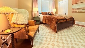 Ein Schlafzimmer mit einem Bett, zwei Nachttischen, einem Sessel, einer Lampe, einem Fenster mit Vorhängen und einem Deckenventilator.
