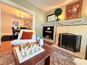 Una sala de estar con chimenea, un tablero de ajedrez, un sillón blanco y una estantería.