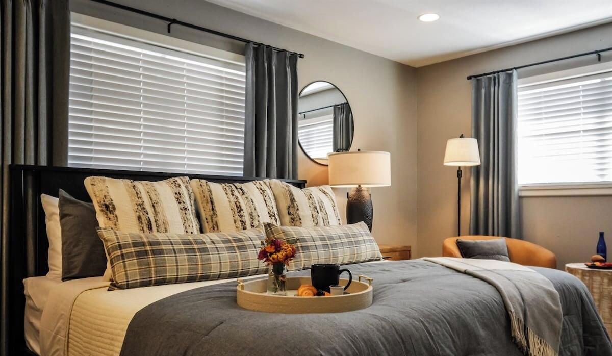 A hotel room with a bed, a bedside table with a lamp, a chair, and a window with curtains.