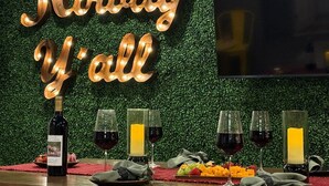 A table with a bottle of wine, glasses, and a candle, set against a backdrop with illuminated text. - Howdy Y’all Ranch | Luxe Western Retreat (Abilene)