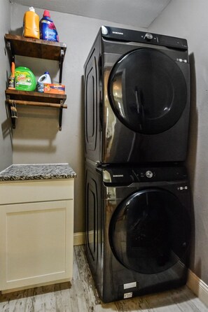 A laundry area with two stacked washers and dryers, a countertop, and shelves with cleaning supplies. - Howdy Y’all Ranch | Luxe Western Retreat (Abilene)