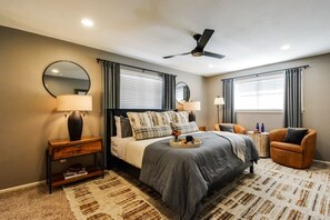 Quarto com cama, mesas de cabeceira, uma cadeira, ventilador de teto e janela com cortinas.
