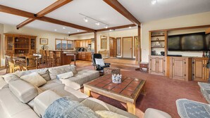 A spacious living room with a large sectional sofa, a coffee table, and a television. There is a kitchen area with wooden cabinets and a dining area with chairs and a table.