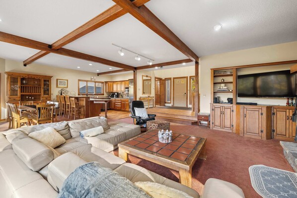 A spacious living room with a large sectional sofa, a coffee table, and a television. There is a kitchen area with wooden cabinets and a dining area with chairs and a table.