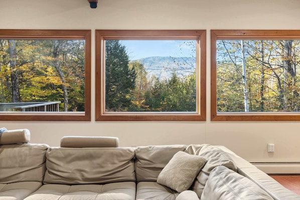 A beige sectional sofa with three windows offering views of autumn foliage and a distant mountain range.