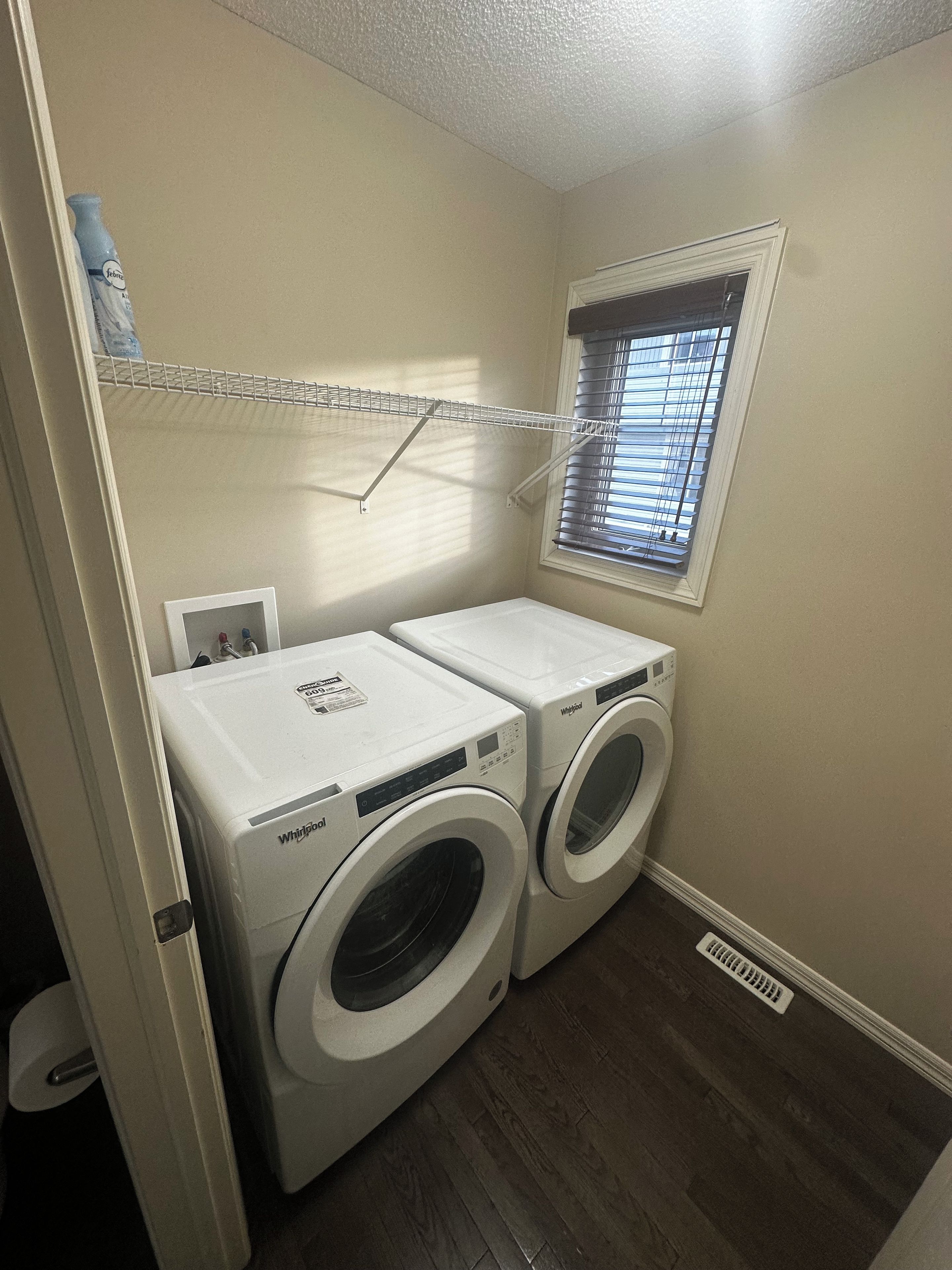 A laundry room with two front-loading washers and dryers, a window with blinds, and a metal shelf above the dryers.
