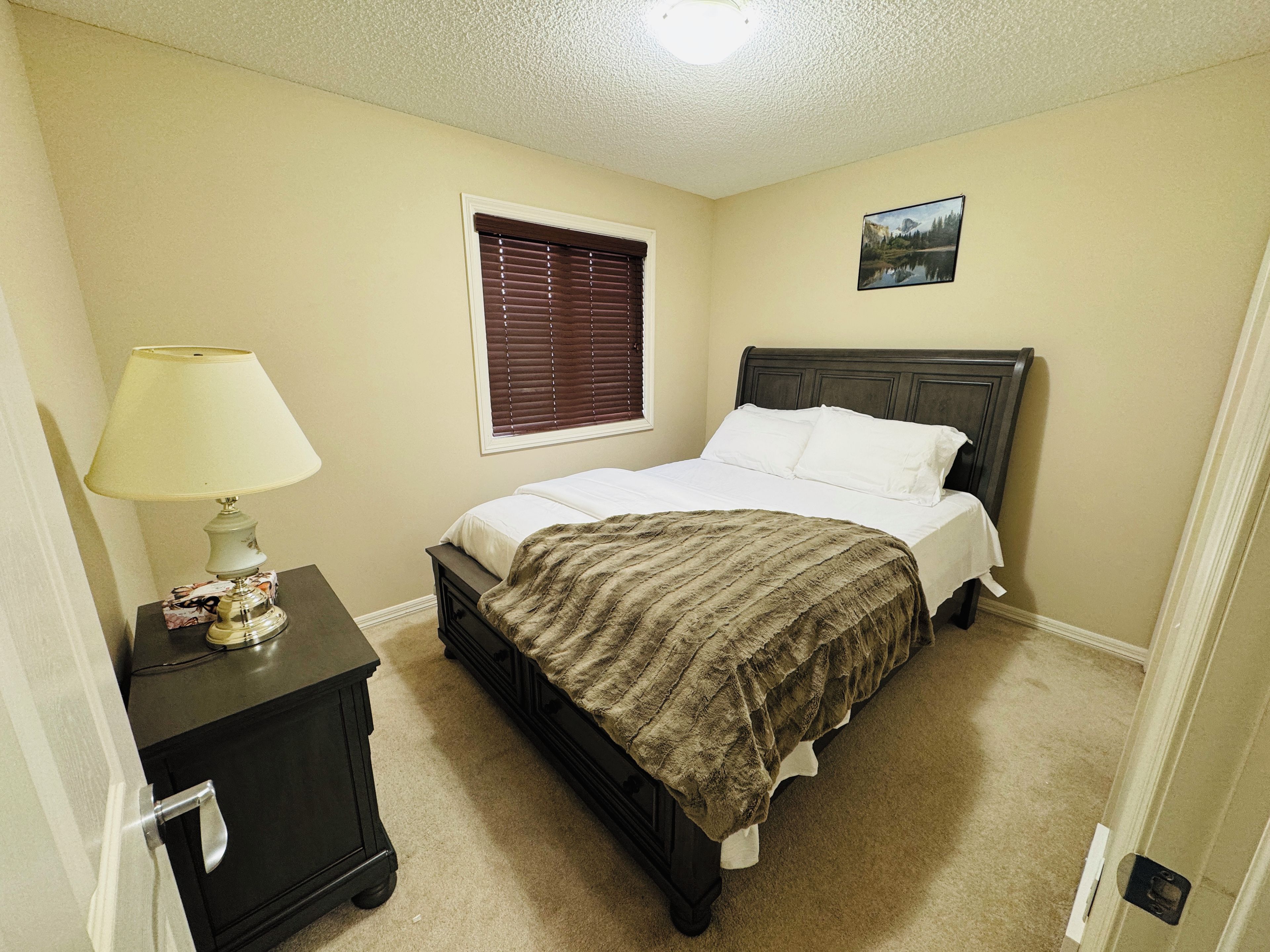 A bedroom with a bed, a nightstand, a lamp, a window with blinds, and a picture on the wall.