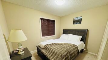 A bedroom with a bed, a nightstand, a lamp, a window with blinds, and a picture on the wall.