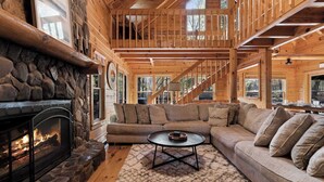 A cozy cabin interior with a stone fireplace, a large sofa, and a staircase leading to an upper level. - Log Cabin by the Lake|Hot Tub, Fire Pit, Games (Blue Ridge)