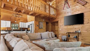 A cozy cabin interior with a large wooden-framed living area, a kitchen with white cabinets, and a mounted flat-screen TV. - Log Cabin by the Lake|Hot Tub, Fire Pit, Games (Blue Ridge)