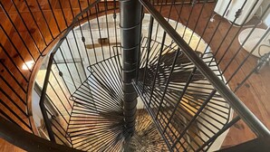 A spiral staircase with a metal railing, wooden floor, and a bookshelf in the background.