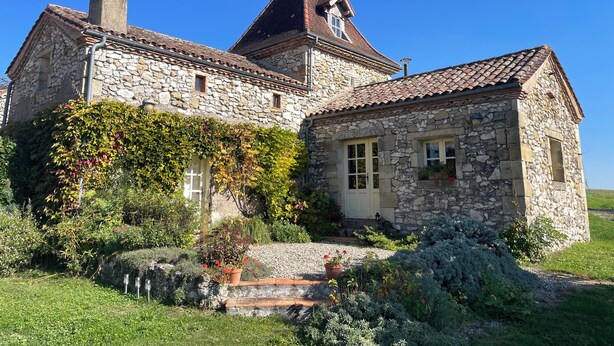 A stone house with a tiled roof, a chimney, and a small garden with potted plants.