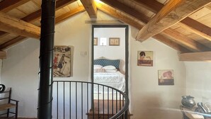 A rustic attic bedroom with a wooden ceiling, a spiral staircase, a bed with a blue headboard, and a wall with vintage posters.