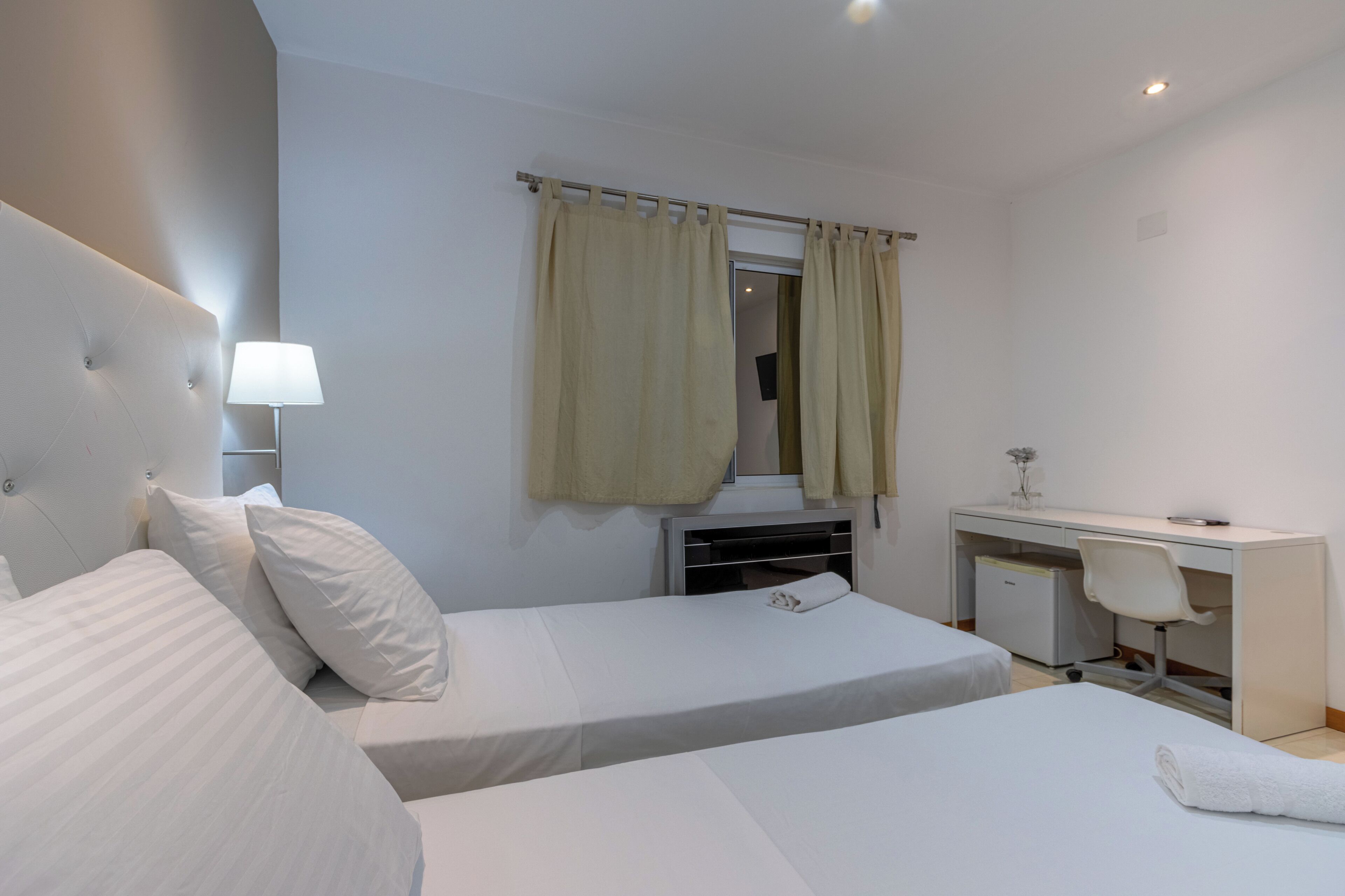 Habitación de hotel con dos camas, un escritorio, una silla, una lámpara y un ventanal con cortinas.