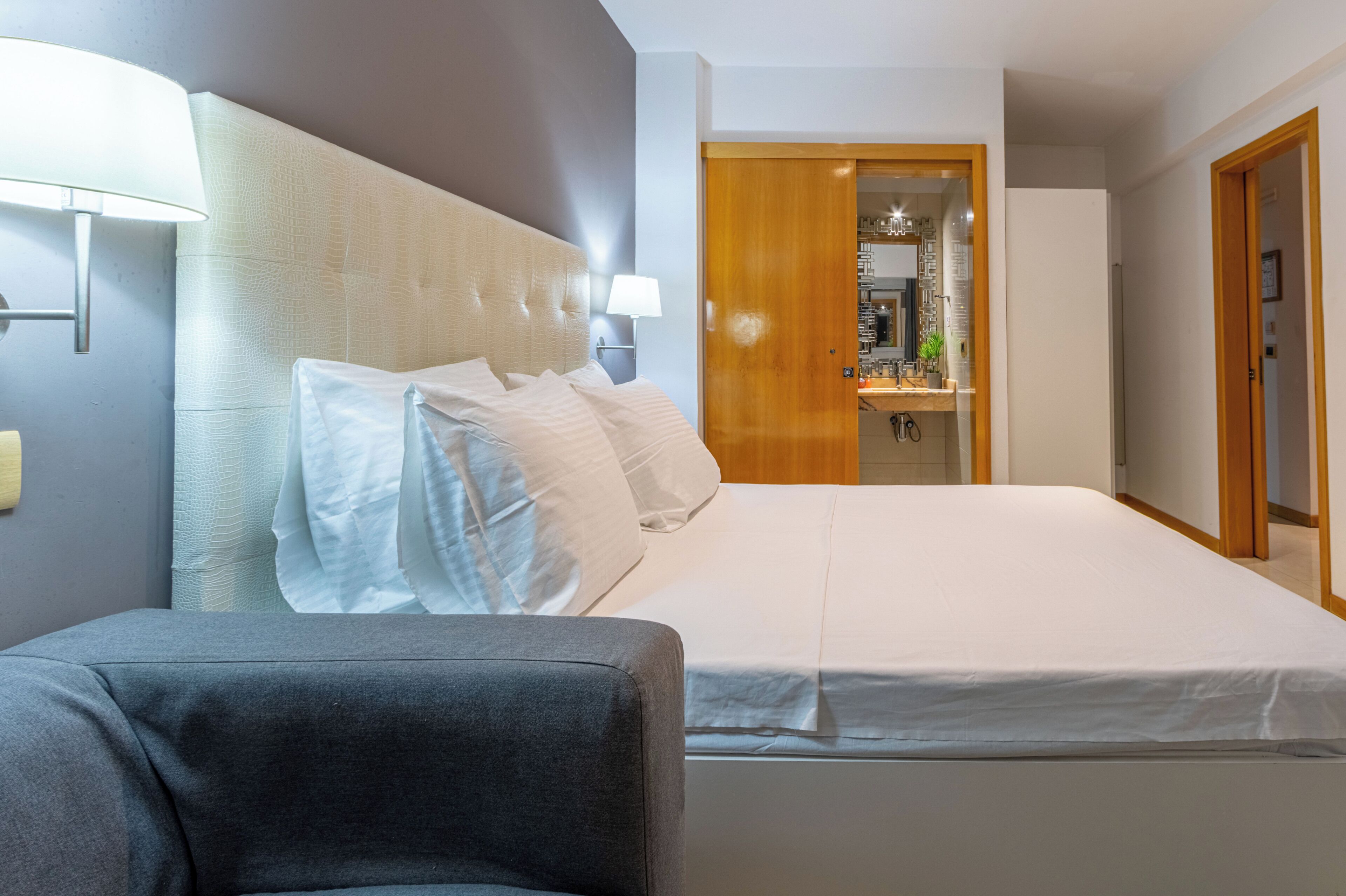 Habitació d'hotel amb un llit, una cadira, una taula de nit, una làmpada i una porta de fusta.