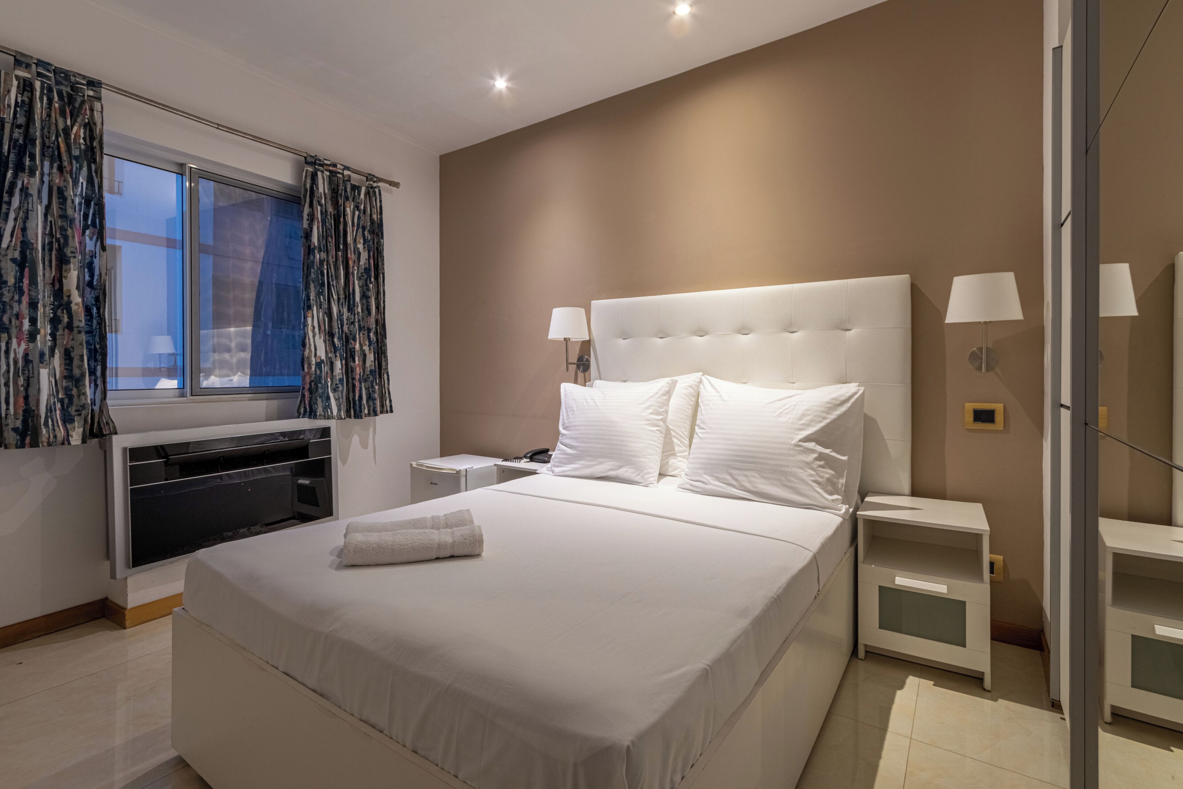 Un dormitorio moderno con una cama grande, mesitas de noche, una lámpara de pared y cortinas en la ventana.