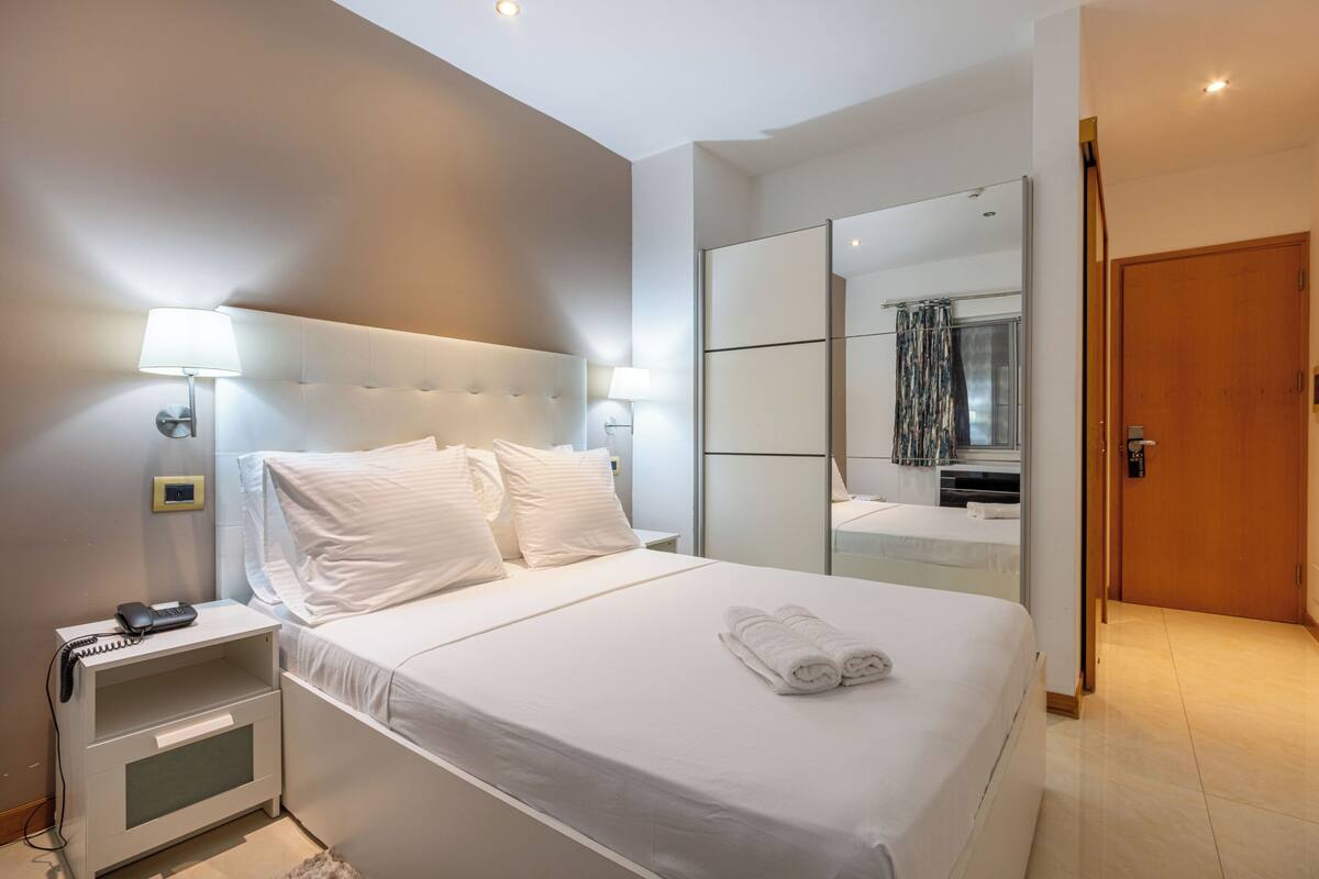 A modern hotel room with a large bed, bedside lamps, a nightstand, a mirror, and a wooden door.