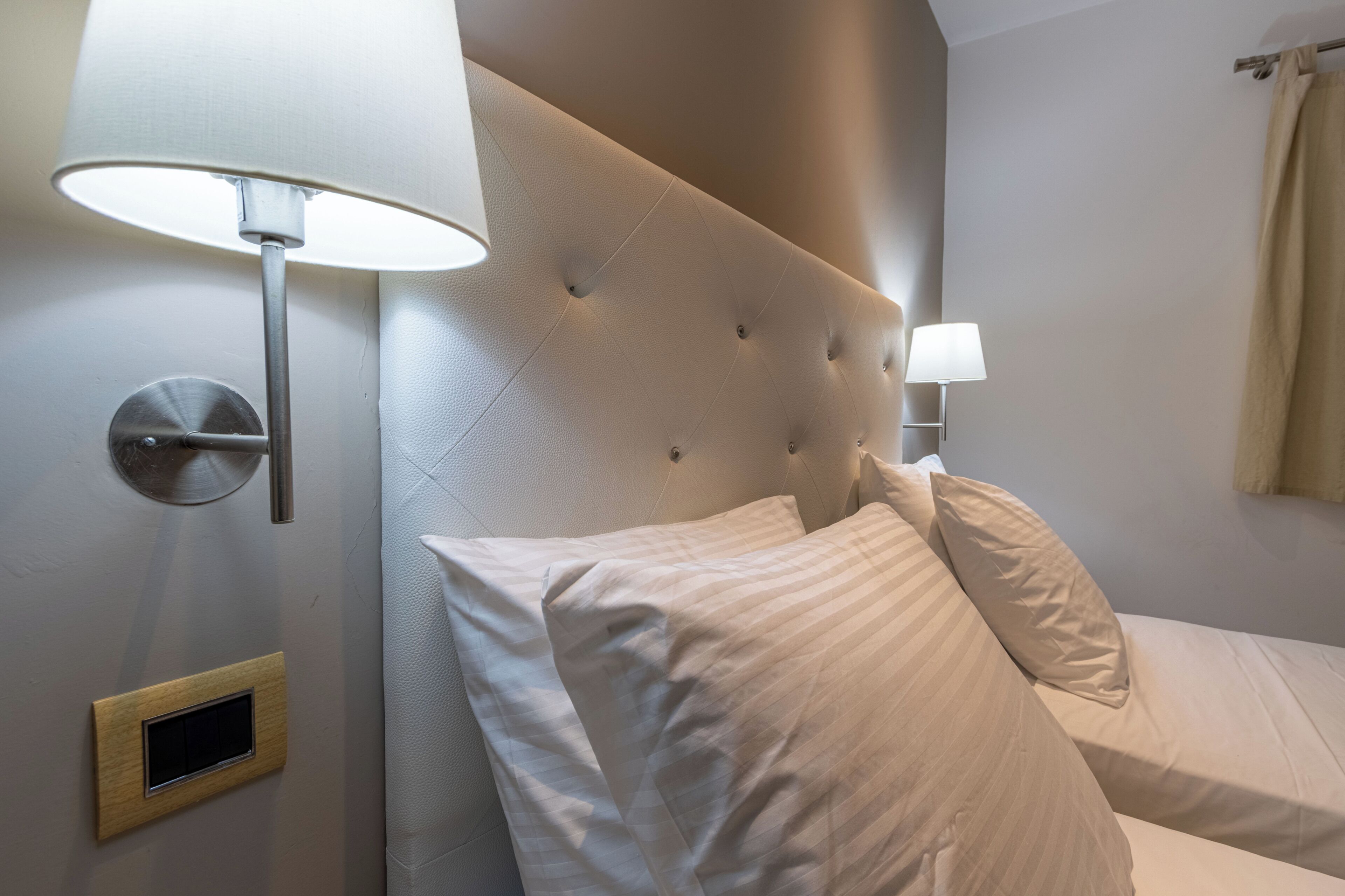 A hotel room with a tufted headboard, two bedside lamps, a wooden switchboard, and a bed with white bedding.