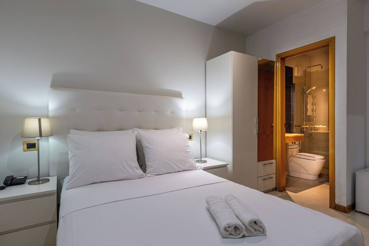 A hotel room with a bed, bedside lamps, a telephone, and a bathroom visible through an open door.
