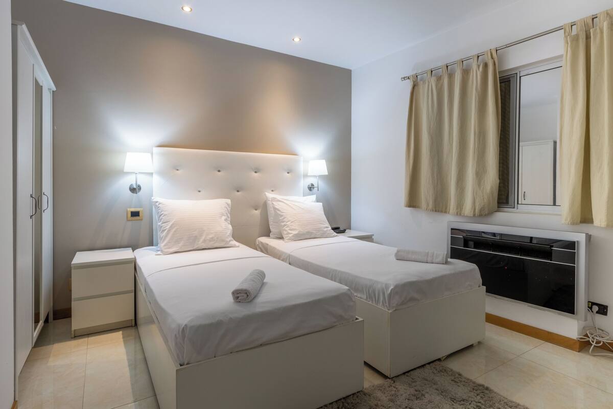 A hotel room with two beds, bedside lamps, a heater, and a window with curtains.