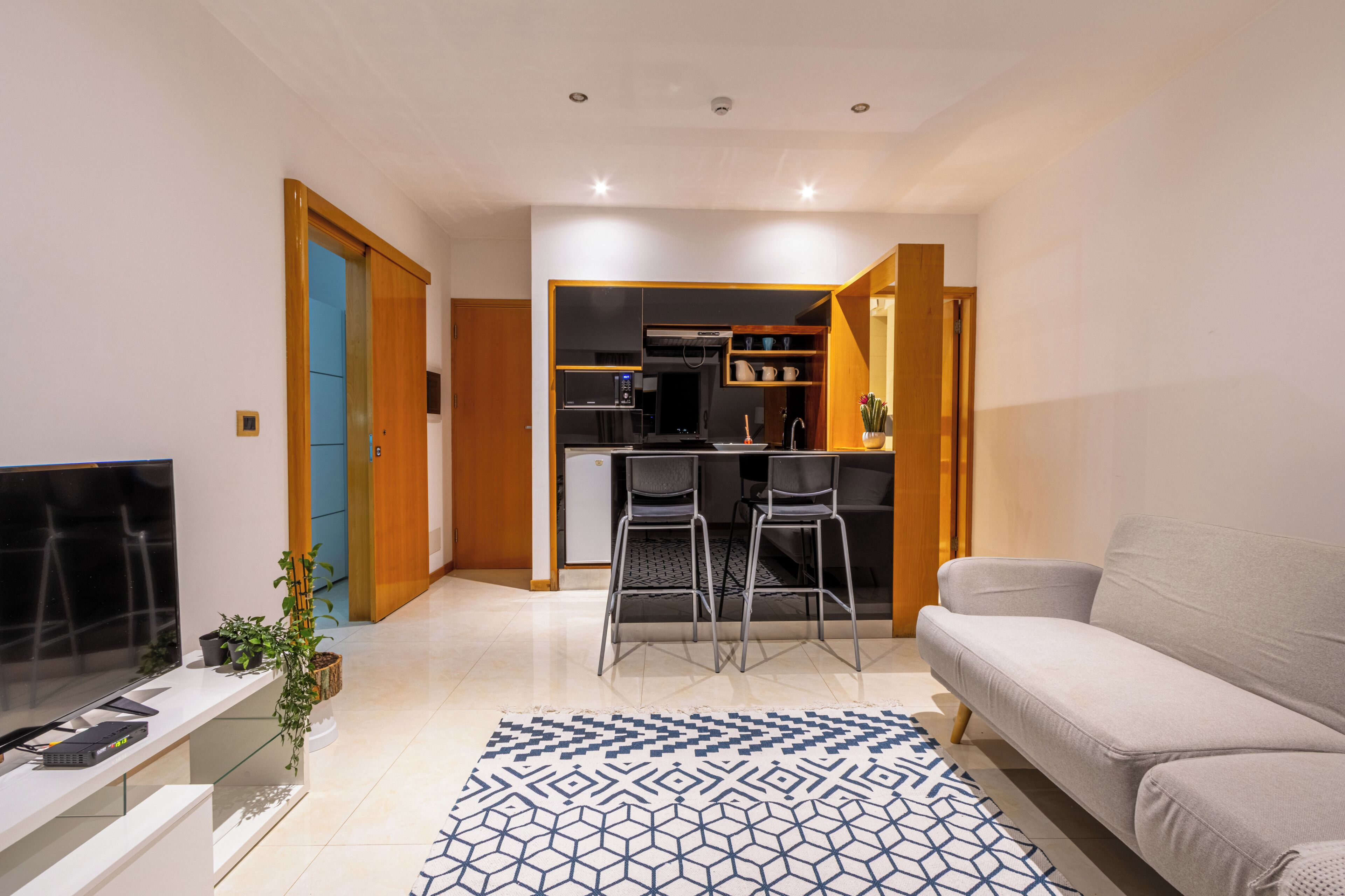 A modern living room with a TV, seating, a patterned rug, and an open kitchen area.