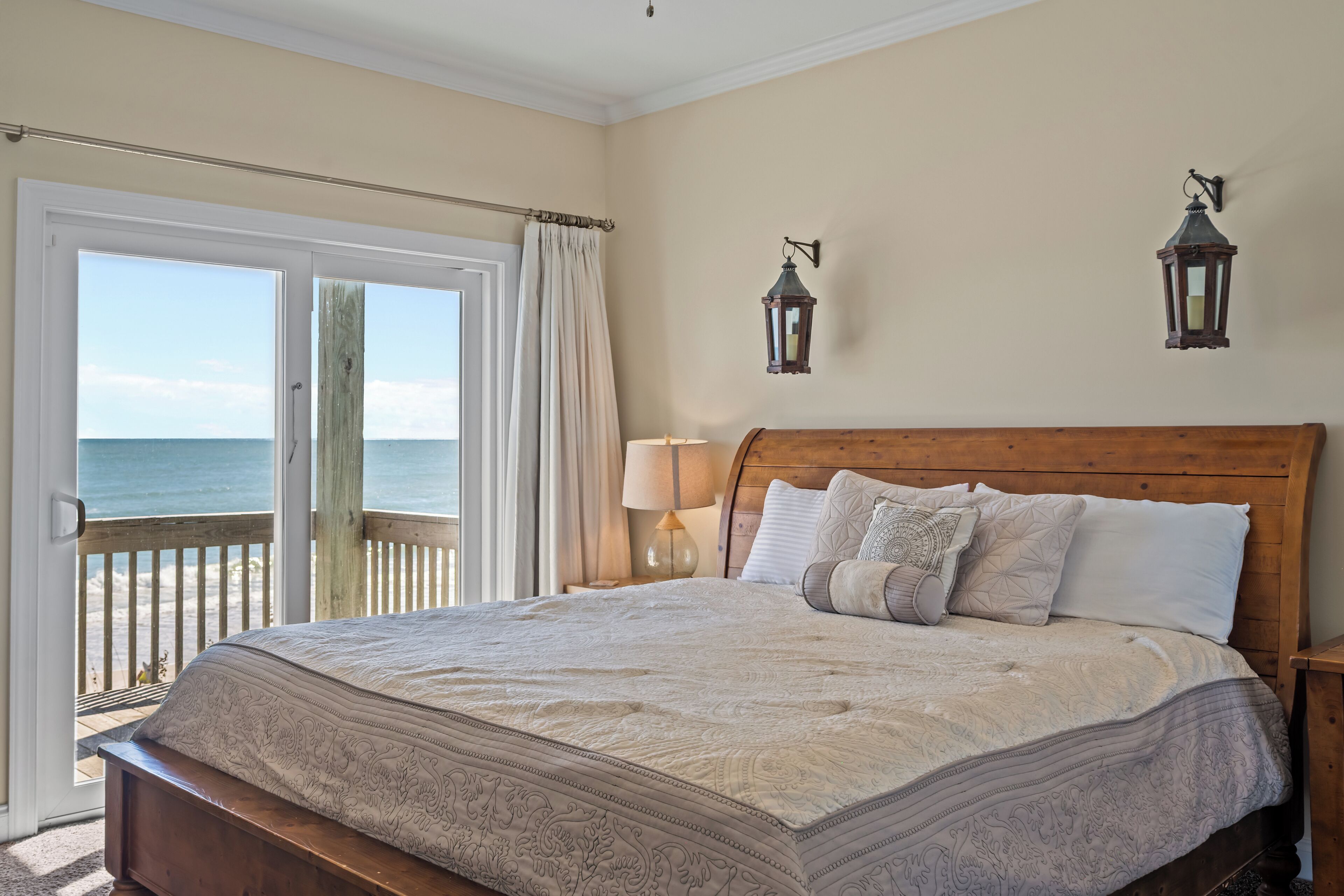 A bedroom with a large bed, a wooden headboard, two wall-mounted lanterns, a nightstand with a lamp, and a view of the ocean through a sliding door.