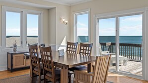 Ein Essbereich mit Holztisch und -stühlen, großen Fenstern mit Meerblick und Blick auf den Strand durch die Schiebeglastür.