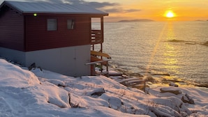 Eine Holzhütte mit einem Balkon mit Blick auf eine verschneite Landschaft und einen Sonnenuntergang über dem Wasser.