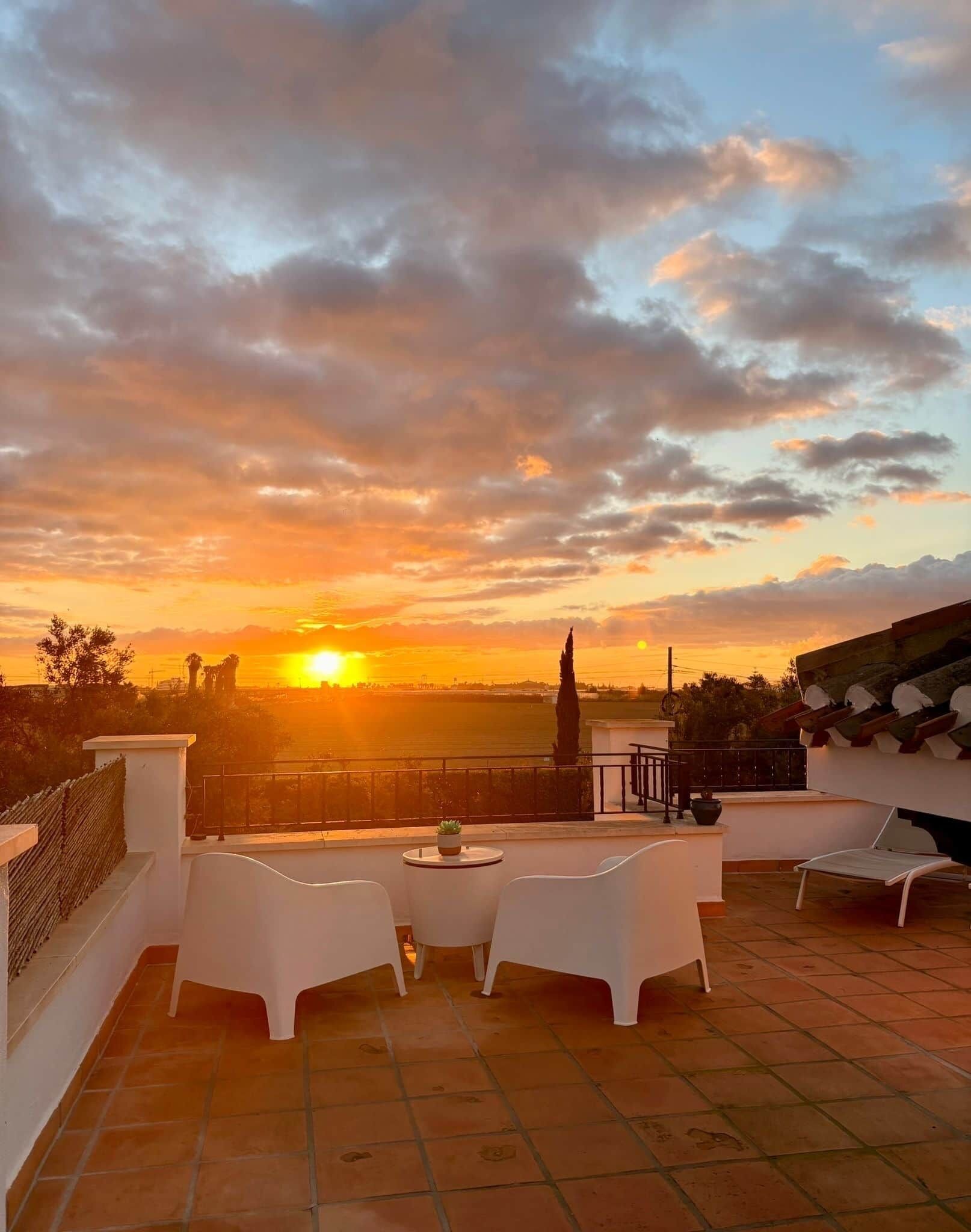 A terrace with white furniture, a potted plant, and a view of the sunset over a landscape.