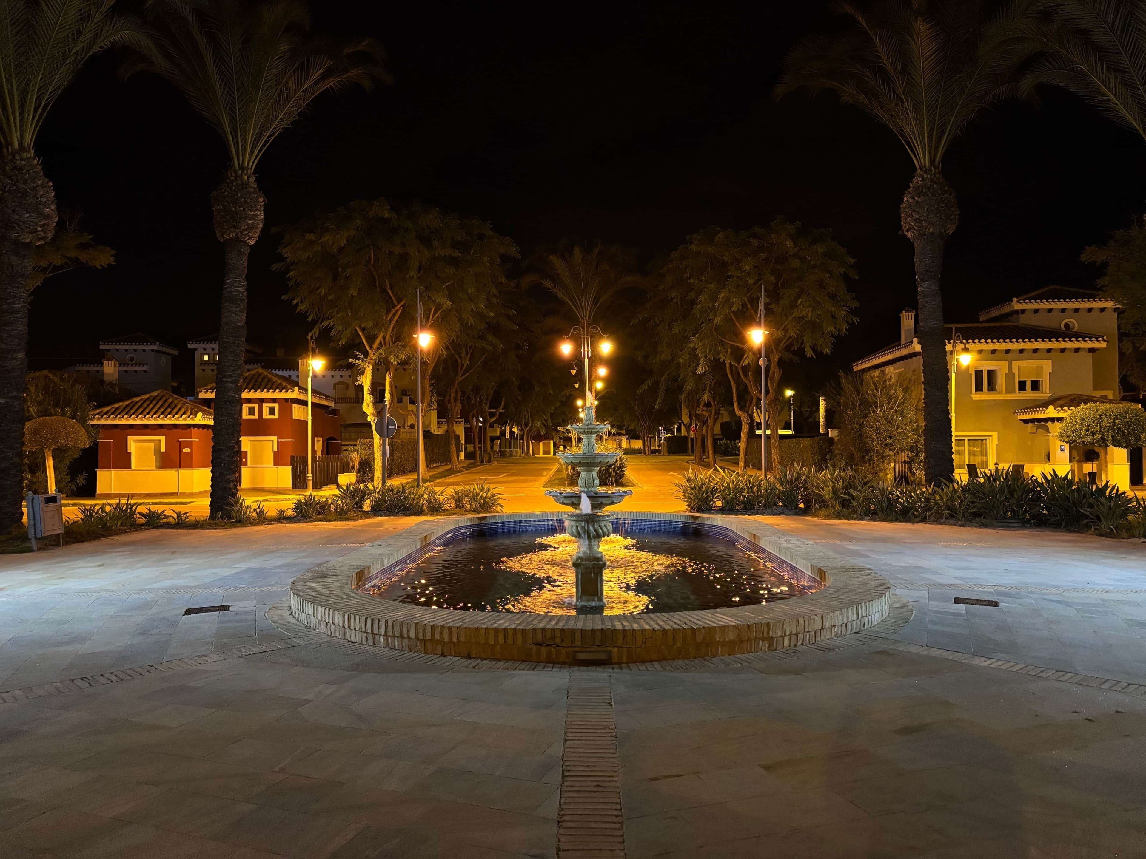 Vista nocturna de una fuente bien iluminada rodeada de palmeras y farolas, con edificios al fondo.
