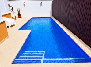 Una piscina interior rectangular con agua azul cristalina, rodeada de suelo con baldosas y una pared de madera oscura.