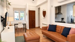 Una sala de estar con un sofá de cuero marrón, una mesa de comedor con sillas, un televisor y una zona de cocina visible a través de un puerta abierta.