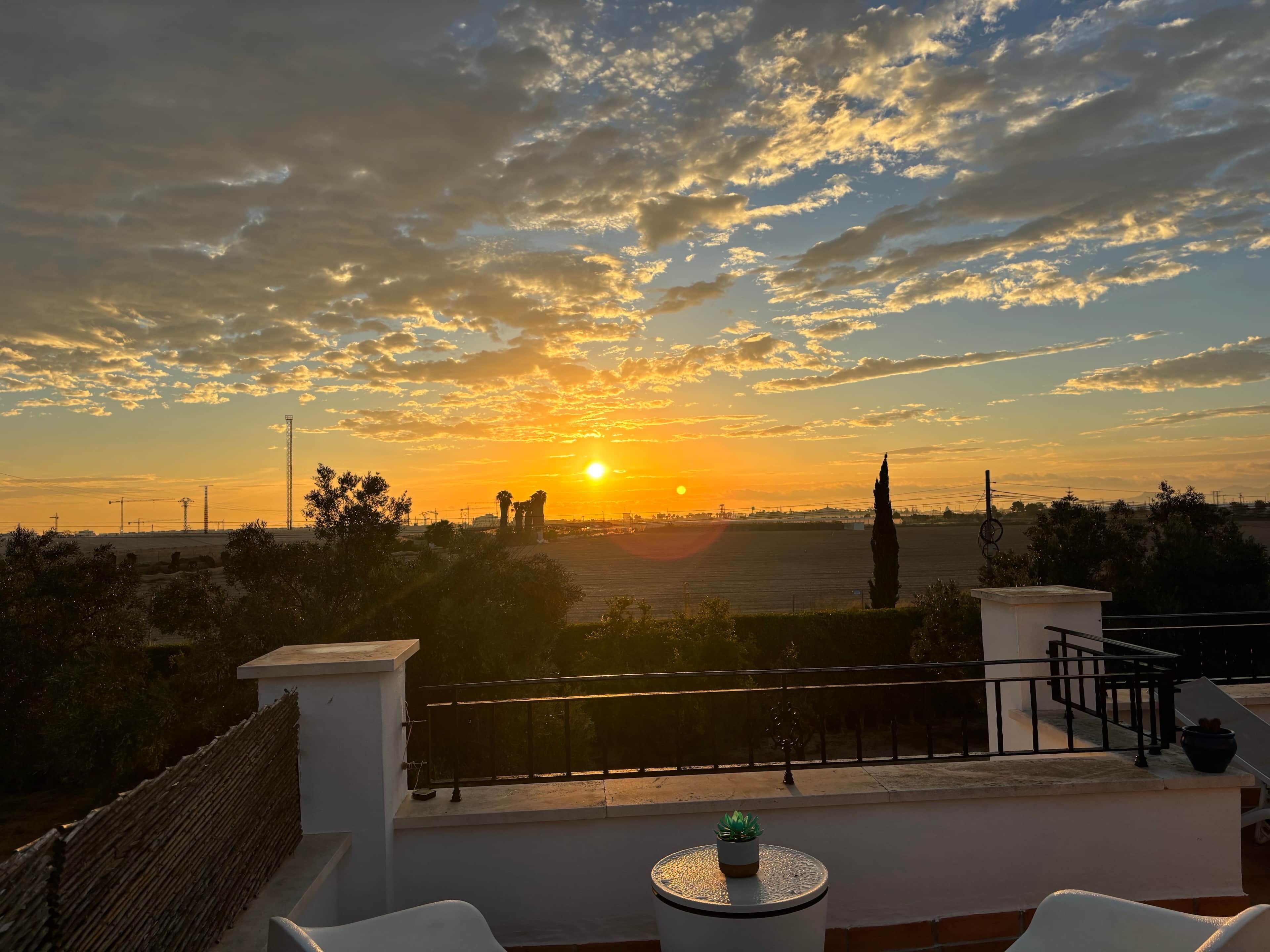 Une vue sur le coucher de soleil depuis un balcon doté d’une balustrade, une petite table avec une plante et un paysage lointain.