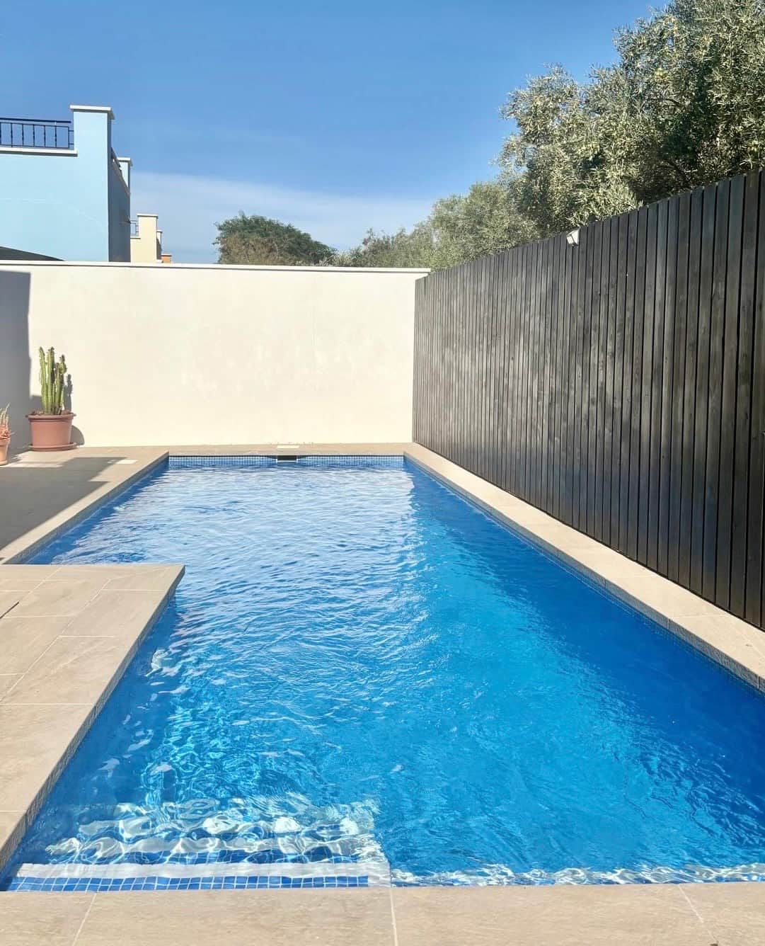 Ein rechteckiger Swimmingpool mit klarem, blauem Wasser, umgeben von einer gefliesten Terrasse und einem Holzzaun.
