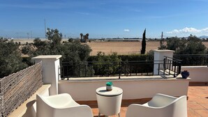 A terrace with two white chairs, a small table with a plant, and a view of a field and distant structures.