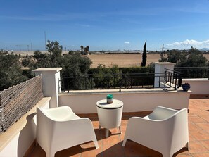A terrace with two white chairs, a small table with a plant, and a view of a field and distant structures. - Villa Alegre, Mar Menor Golf (Torre-Pacheco)
