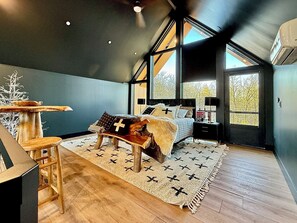 A bedroom with a bed, a nightstand, a window, and a view of trees. - The Lofts @ Frost Fire- Cabin B (Walhalla)