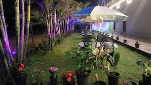 An outdoor seating area with tables and chairs, surrounded by potted plants and bamboo trees, under a brightly lit building. - Hotel Golden Galaxy (Mahabaleshwar)